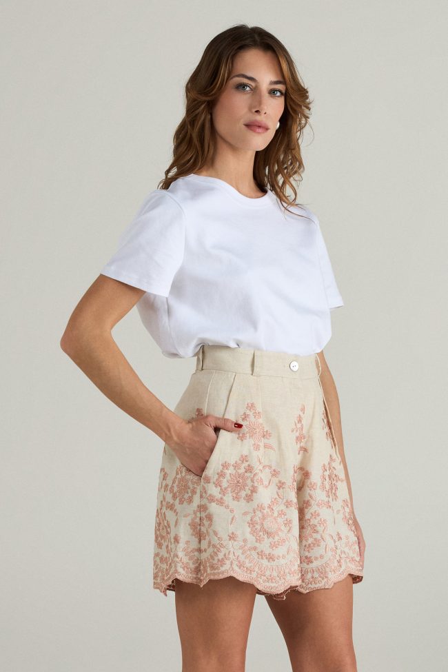 Model wearing high-waisted blush embroidered linen shorts with scalloped hem, styled with a white t-shirt and espadrilles.