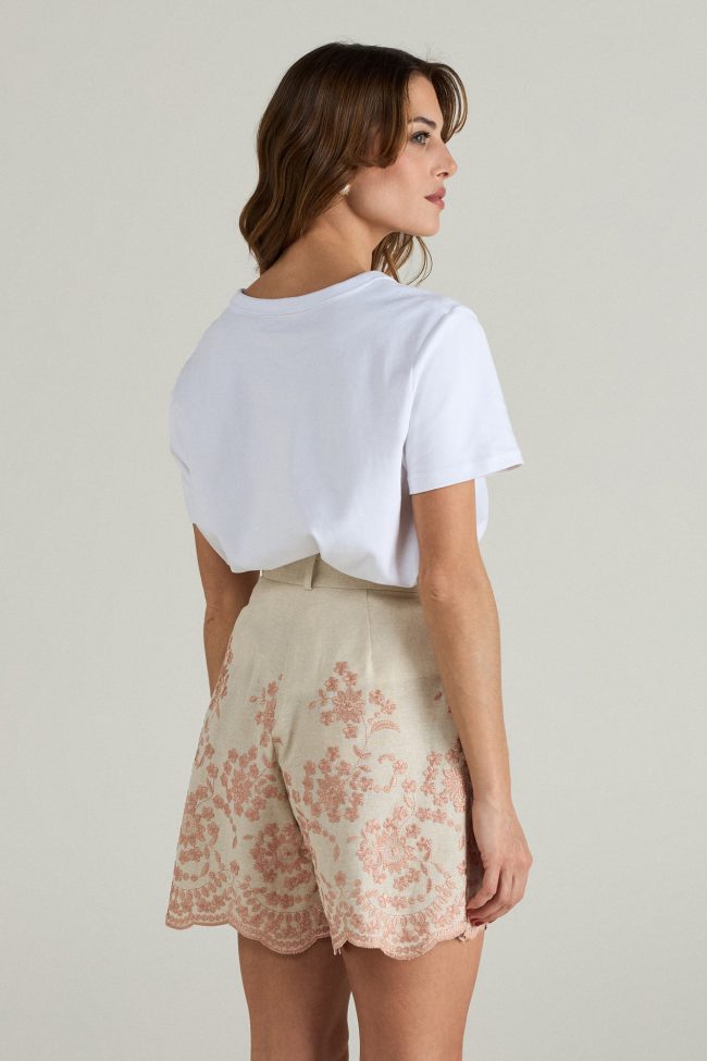 Model wearing high-waisted blush embroidered linen shorts with scalloped hem, styled with a white t-shirt and espadrilles.