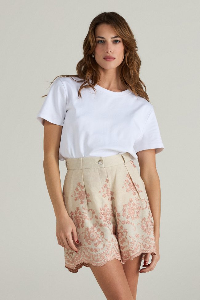 Model wearing high-waisted blush embroidered linen shorts with scalloped hem, styled with a white t-shirt and espadrilles.