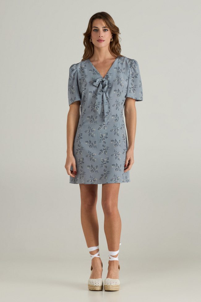 Model wearing a light blue mini dress with short puff sleeves, delicate floral print, and front bow detail.