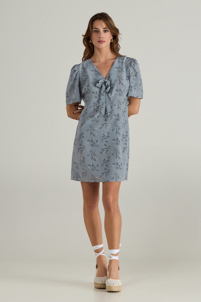 Model wearing a light blue mini dress with short puff sleeves, delicate floral print, and front bow detail.