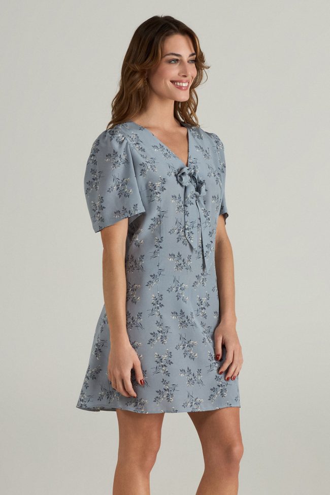 Model wearing a light blue mini dress with short puff sleeves, delicate floral print, and front bow detail.