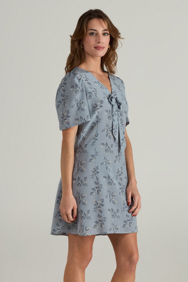 Model wearing a light blue mini dress with short puff sleeves, delicate floral print, and front bow detail.