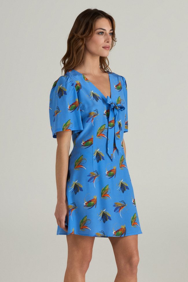 Model wearing blue mini dress with colourful insect print, puff sleeves and a bow tie neckline.